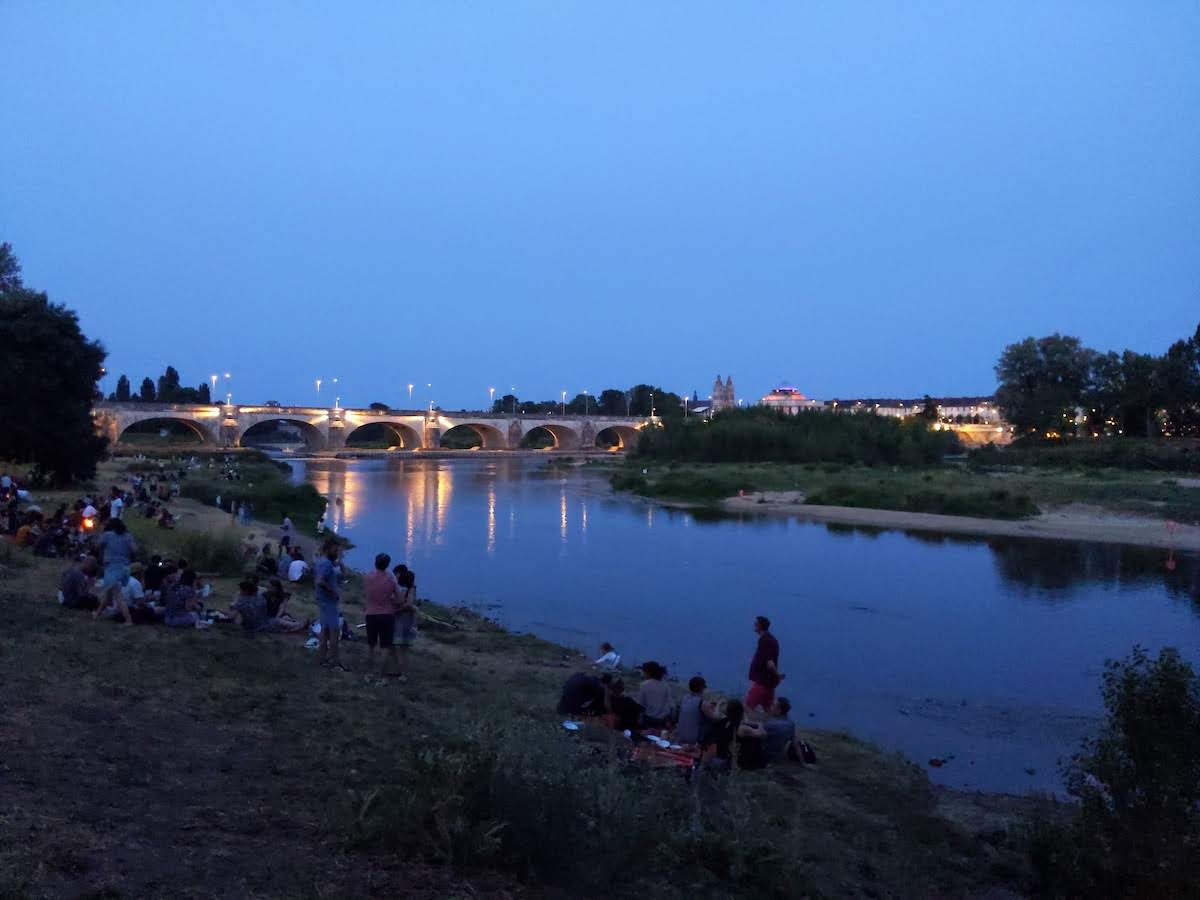 Des gens sont installés en petits groupes sur le bord de Loire pour profiter de la soirée d'été. Plus loin, les lumières d'un pont se reflètent dans la Loire alors que le soleil se couche.
